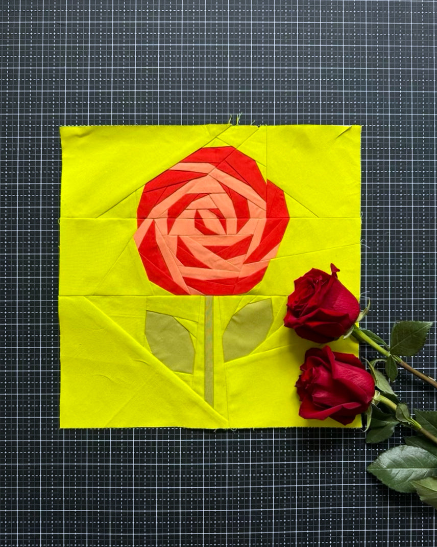 Quilt block with a red rose design on a yellow background, accompanied by real red roses on a grid background.