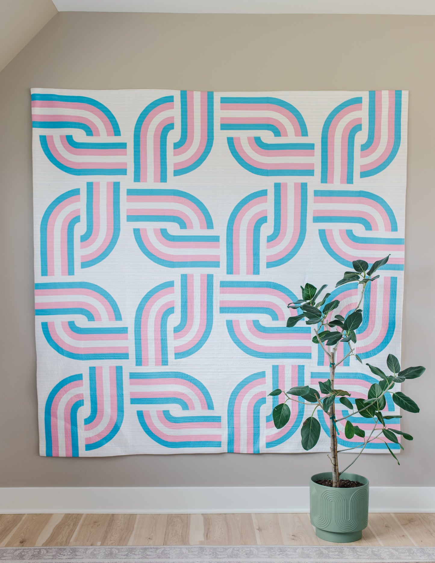 Geometric patterned quilt with pink and blue design on a wall next to a potted plant.