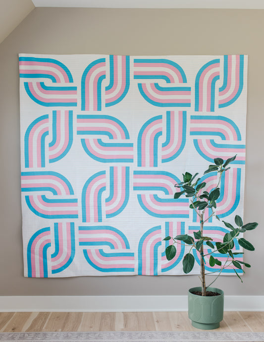 Geometric patterned quilt with pink and blue design on a wall next to a potted plant.