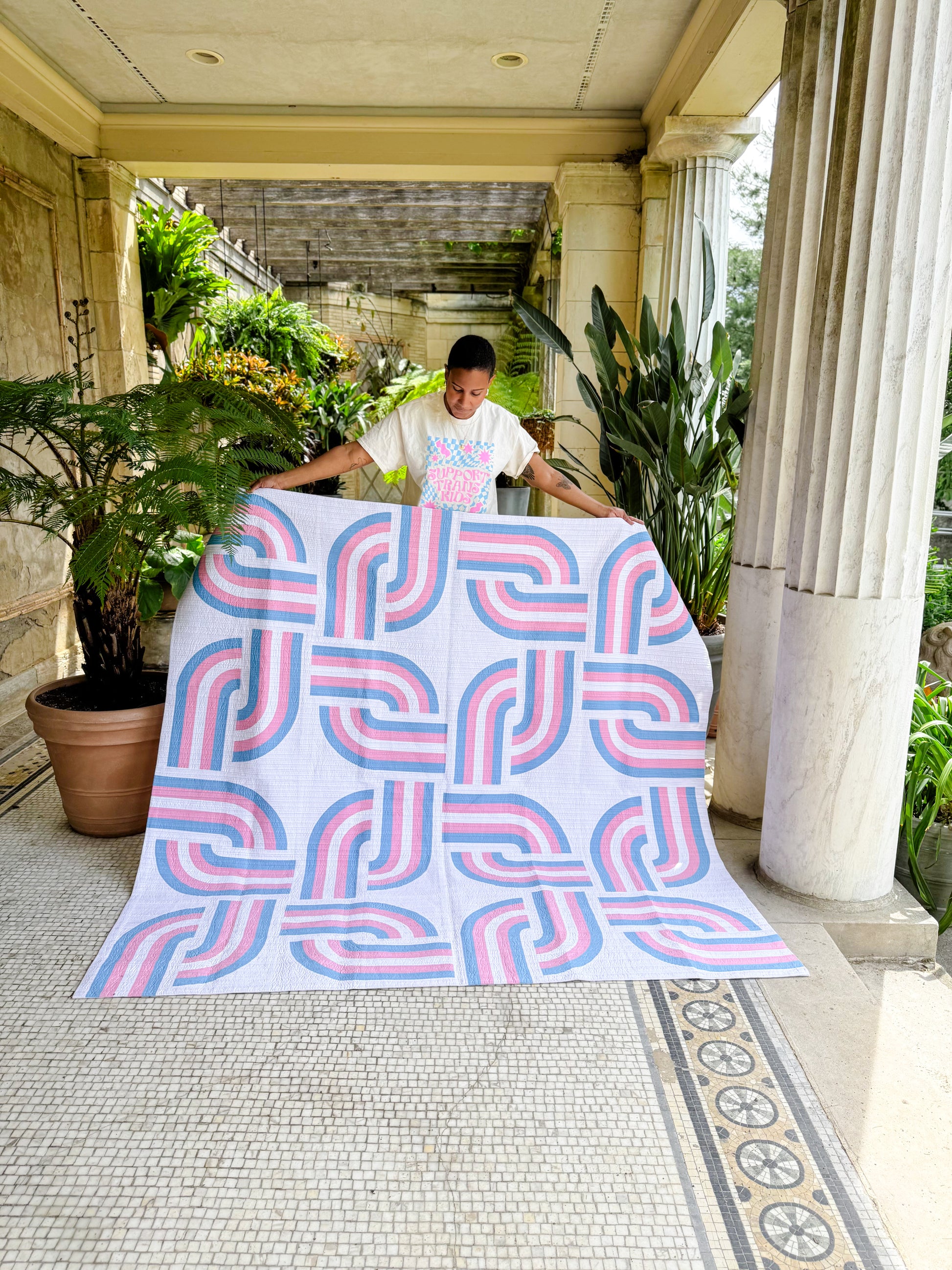 Person holding a large quilt with a pink and blue pattern in an outdoor setting.