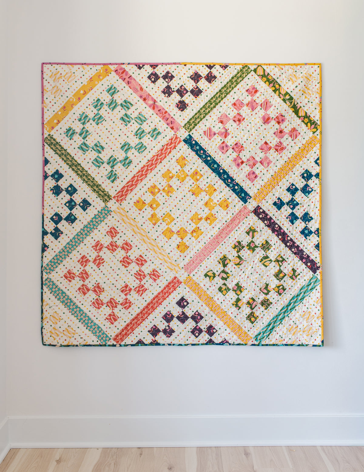 A flat-lay view of the completed "Playdate" quilt hanging against a clean white wall above light-colored hardwood flooring. The quilt features patterned, colorful fabrics set on a cream background with fine polka dots. Diagonal patterned sashing in shades of yellow, green, pink, teal, and orange divides the design into diamond sections containing stepped, pixelated diamond shapes in matching colors.