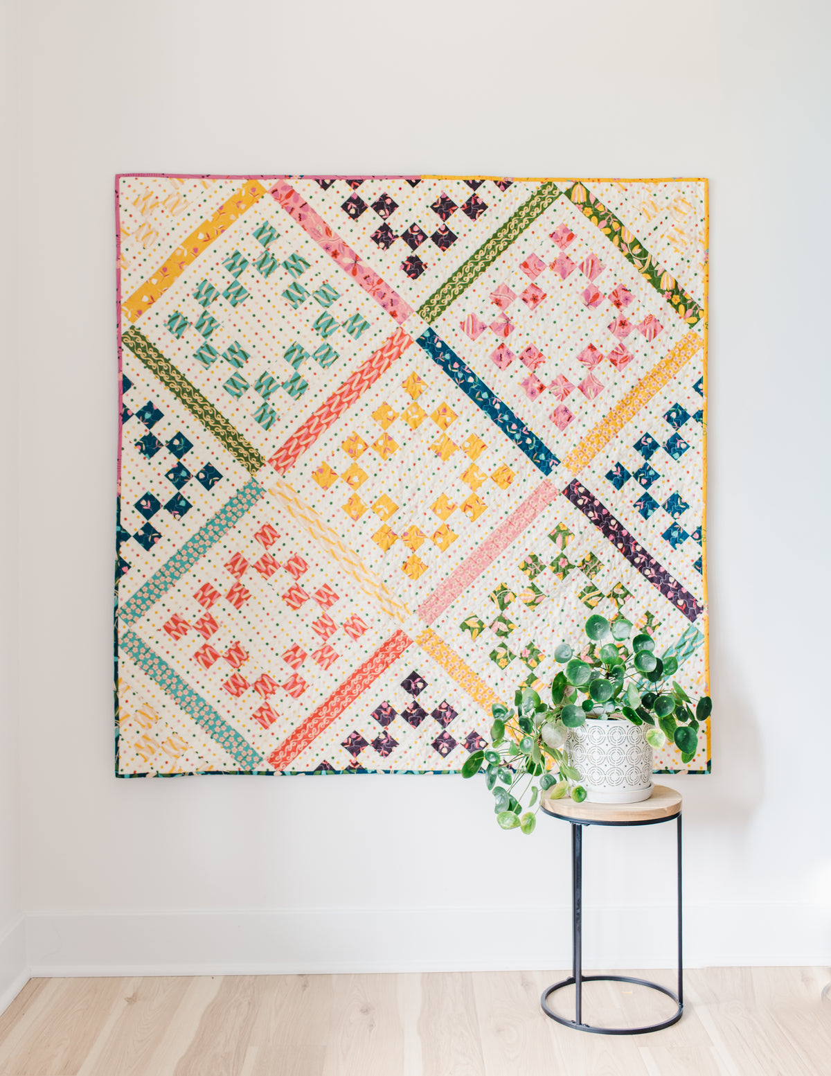 A flat-lay view of the completed "Playdate" quilt hanging against a clean white wall above light-colored hardwood flooring. The quilt features patterned, colorful fabrics set on a cream background with fine polka dots. Diagonal patterned sashing in shades of yellow, green, pink, teal, and orange divides the design into diamond sections containing stepped, pixelated diamond shapes in matching colors. In the bottom-right foreground sits a small round side table with a black metal frame, holding a potted Pilea