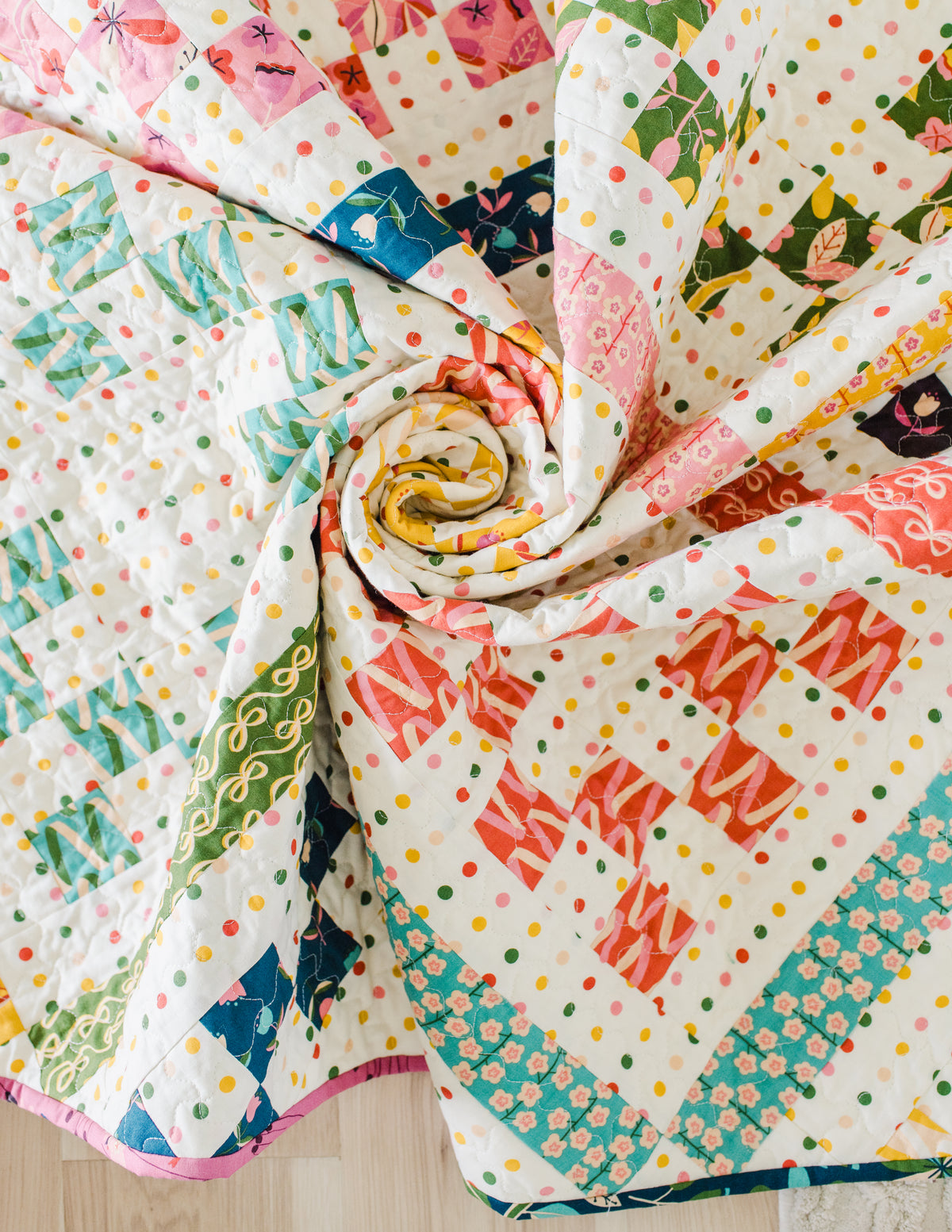 A close-up view of the completed "Playdate" quilt artfully swirled into a spiral pattern in the center. The photo highlights the detailed texture of the quilting stitches, the multicolored polka dot background fabric, and the vibrant floral, ribbon, and geometric fabric prints in shades of pink, yellow, teal, orange, and olive green.