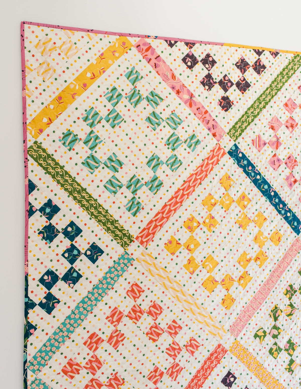A close-up view of the completed "Playdate" quilt hanging against a clean white wall above light-colored hardwood flooring. The quilt features patterned, colorful fabrics set on a cream background with fine polka dots. Diagonal patterned sashing in shades of yellow, green, pink, teal, and orange divides the design into diamond sections containing stepped, pixelated diamond shapes in matching colors. 